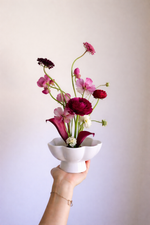 Hand holding a white vase with pink and red flowers against a plain background