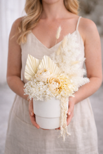 Woman holding a modern white everlasting flower arrangement in a ceramic vase, made with preserved hydrangea, dried palms and delicate textured stems